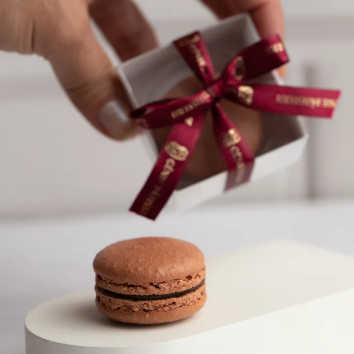 Mão segurando uma caixinha de macaron com laço, como quem oferece um presente.